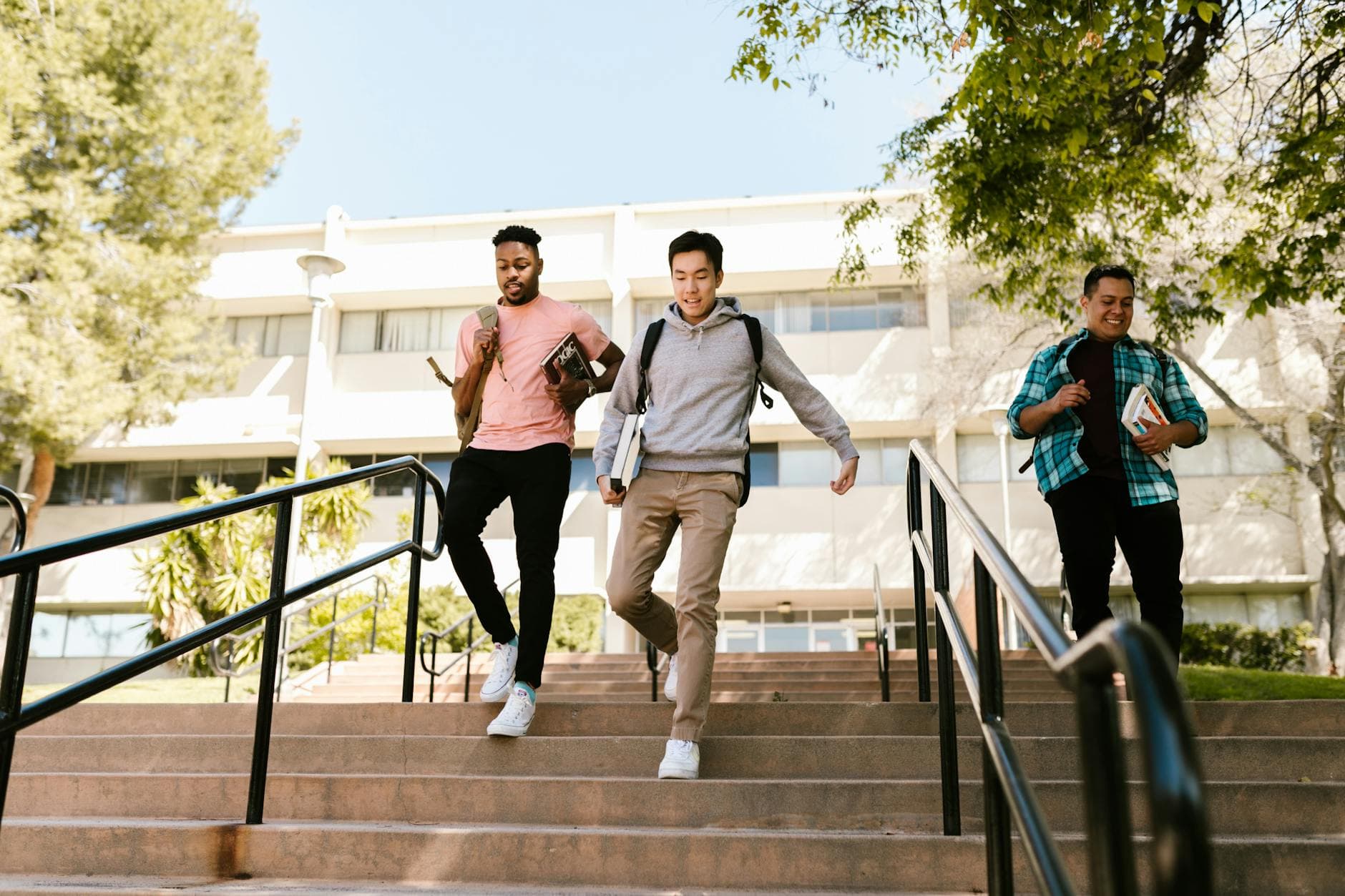 Three college students walk down stairs on campus, carrying books and backpacks, on a sunny day.