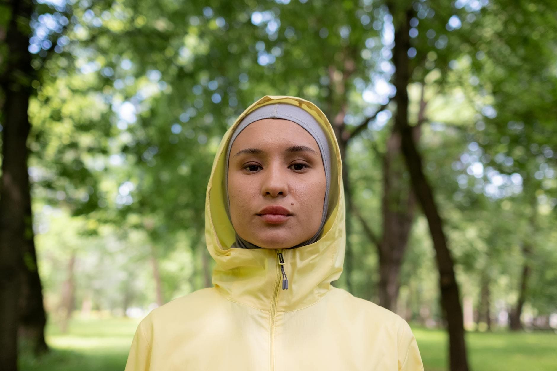 A serene portrait of a woman wearing a hijab and yellow windbreaker in a lush green park.
