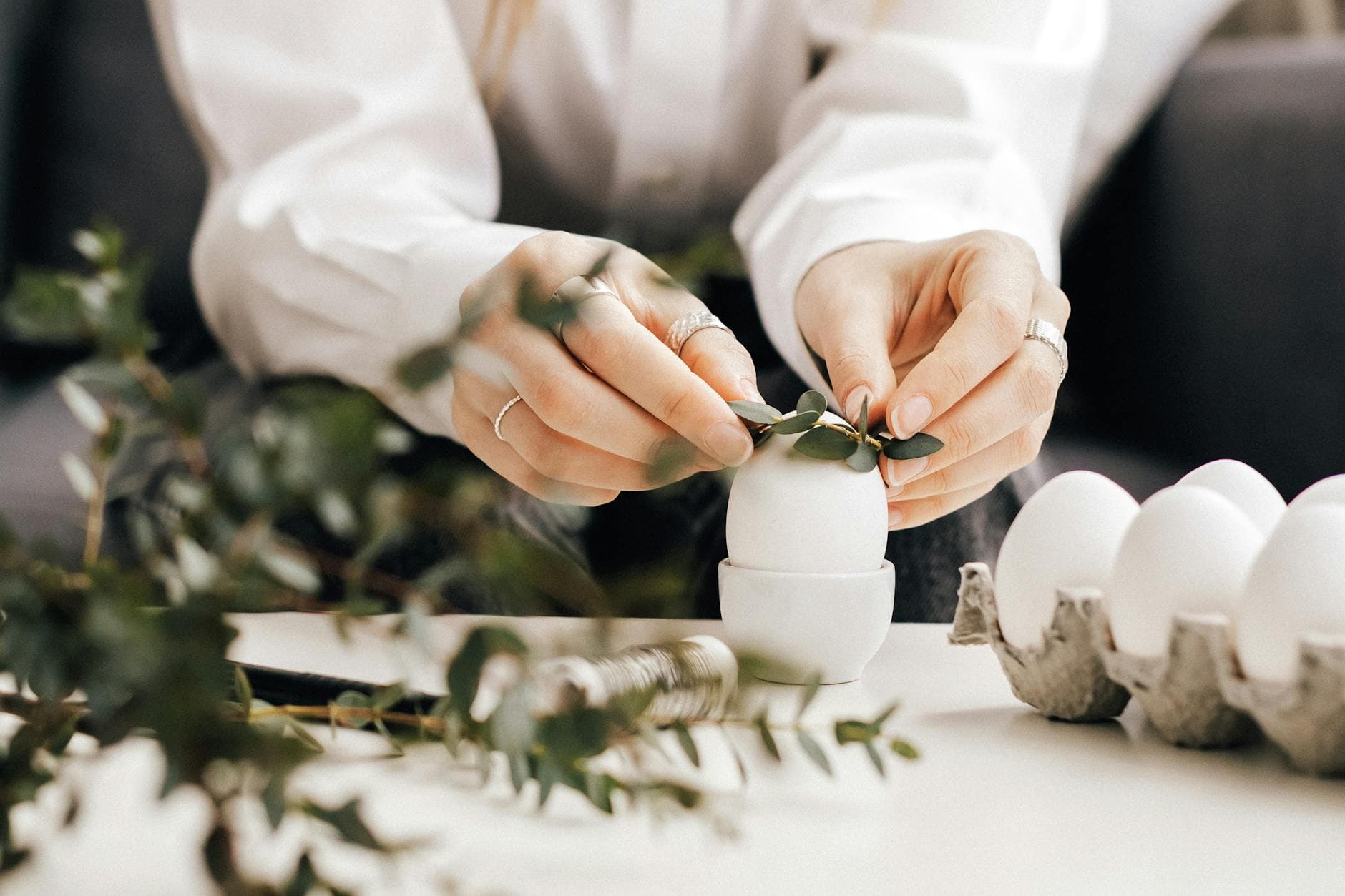 Hands arranging leaves on white eggs for an Easter decoration indoors, creating a natural festive look.
