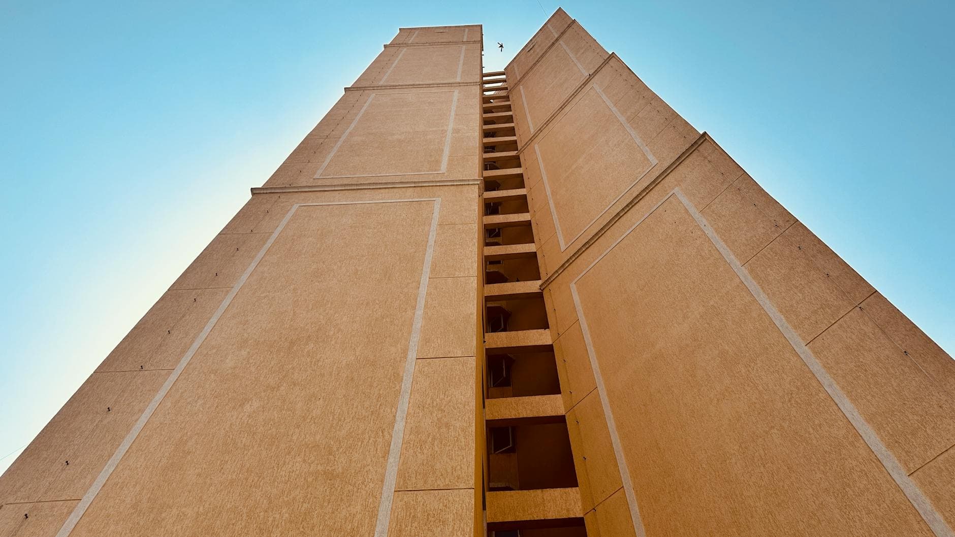 Looking up at a tall beige building against a clear blue sky, showcasing urban architecture.