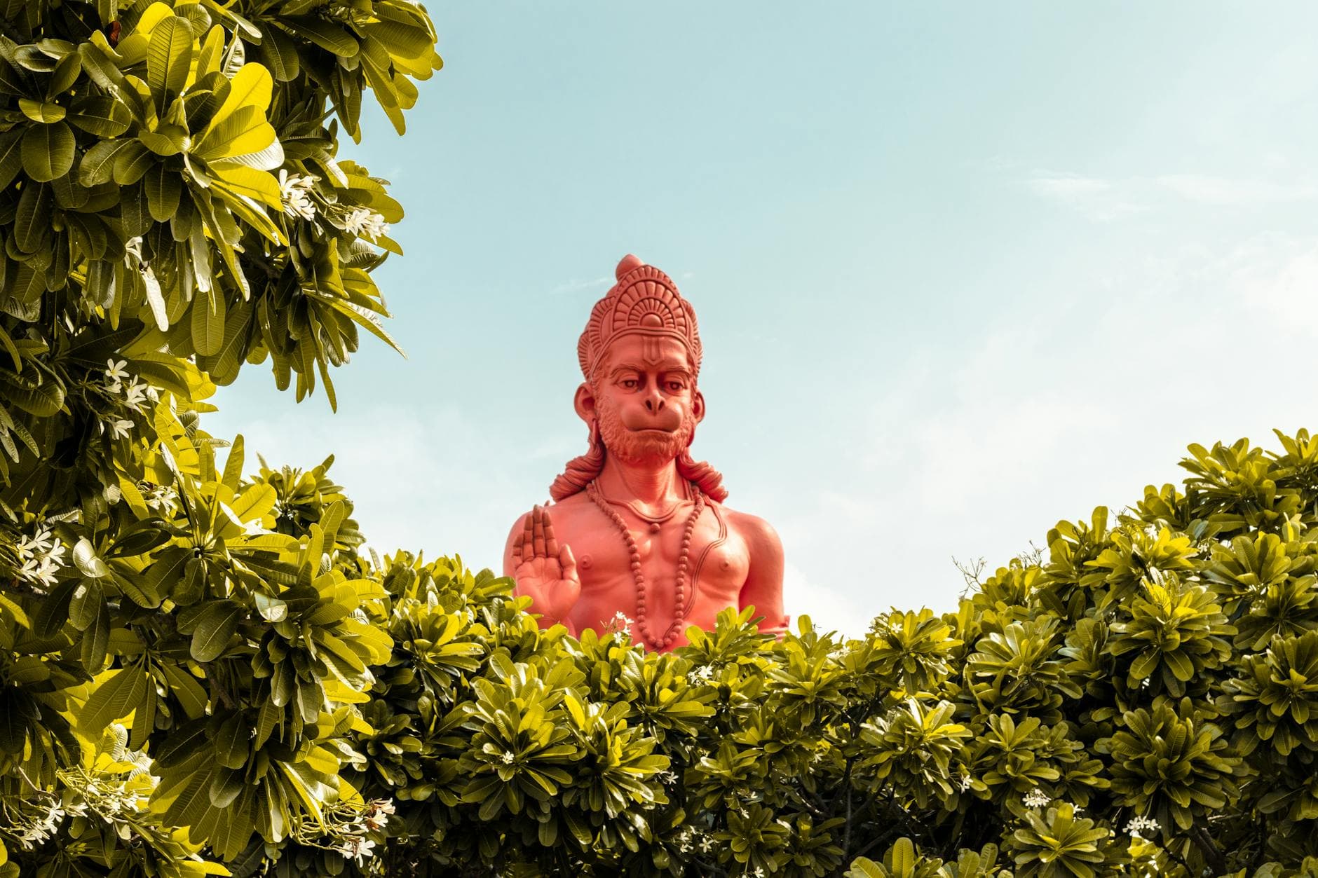 A vibrant red statue of Hanuman surrounded by green foliage under a clear sky.