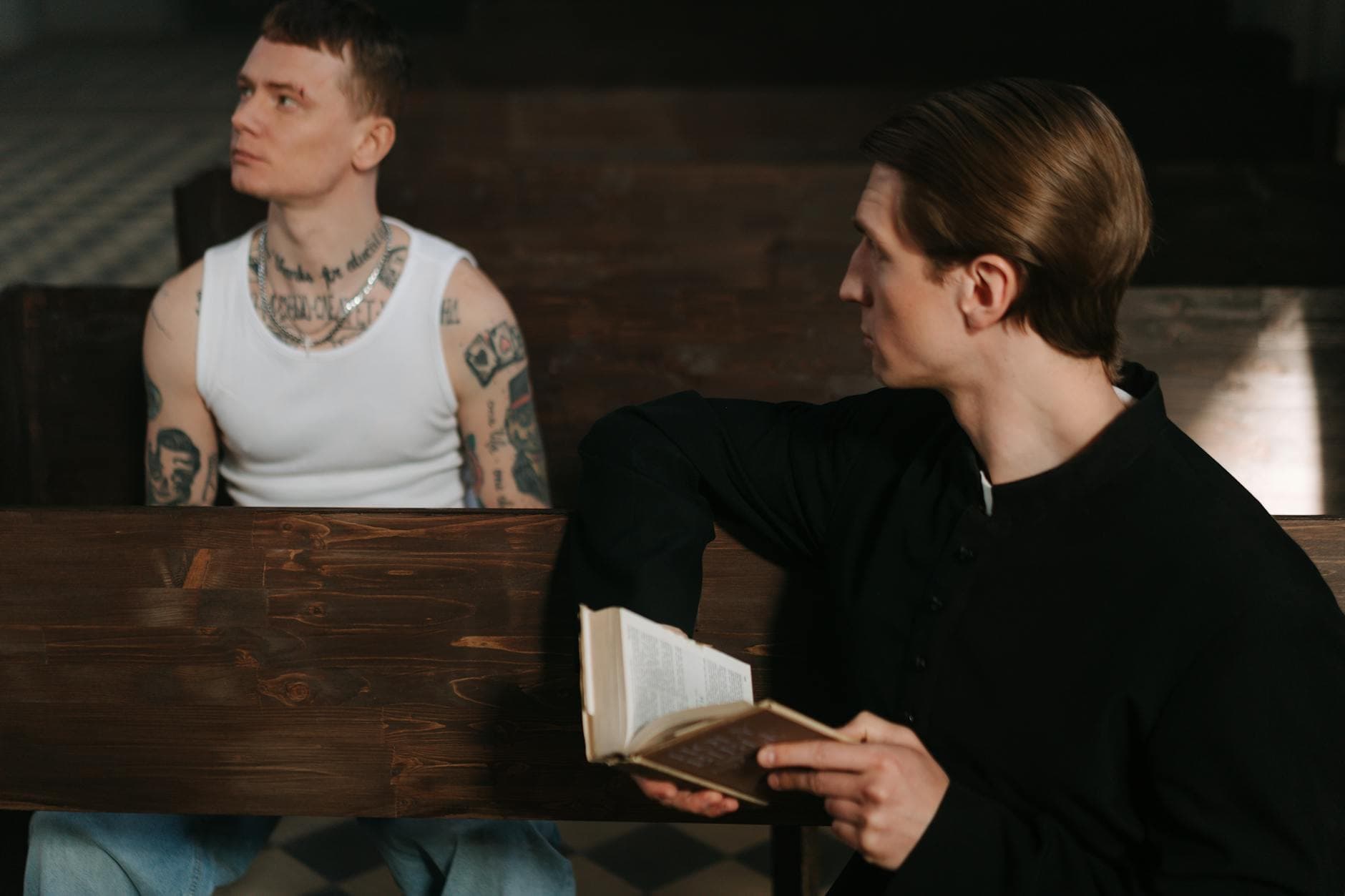 A tattooed man in a white tank top sits opposite a priest holding a holy book, symbolizing a contrast of lifestyles and beliefs.