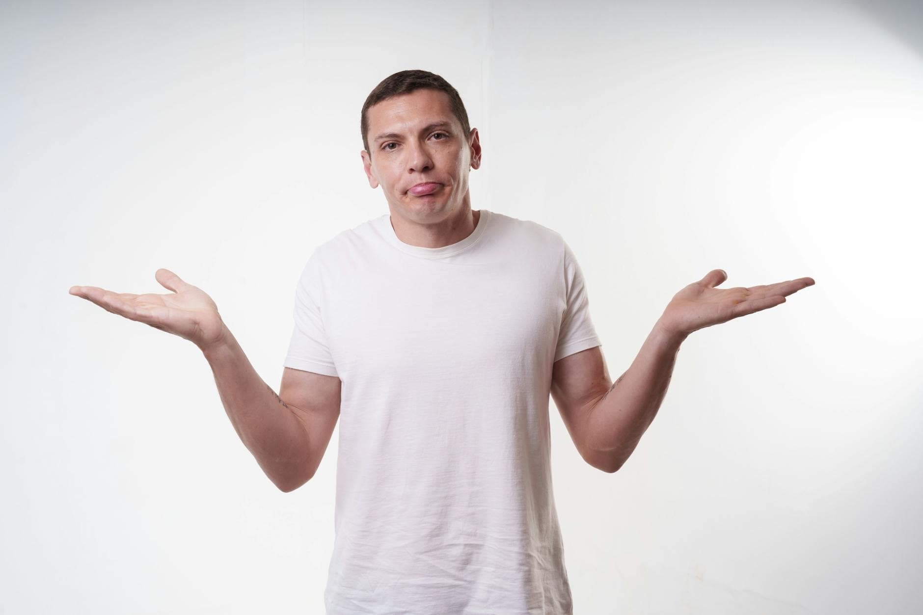 Adult man in white t-shirt shrugs in studio, expressing confusion against a plain background.