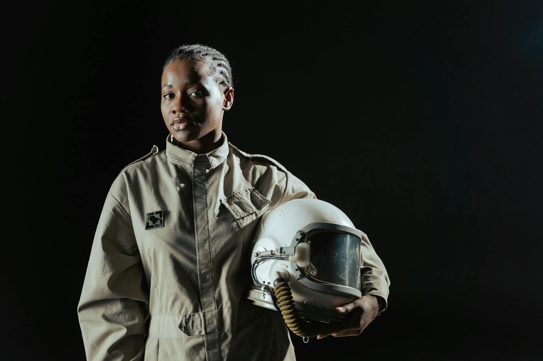 A confident female astronaut in a spacesuit holding her helmet against a dark background.