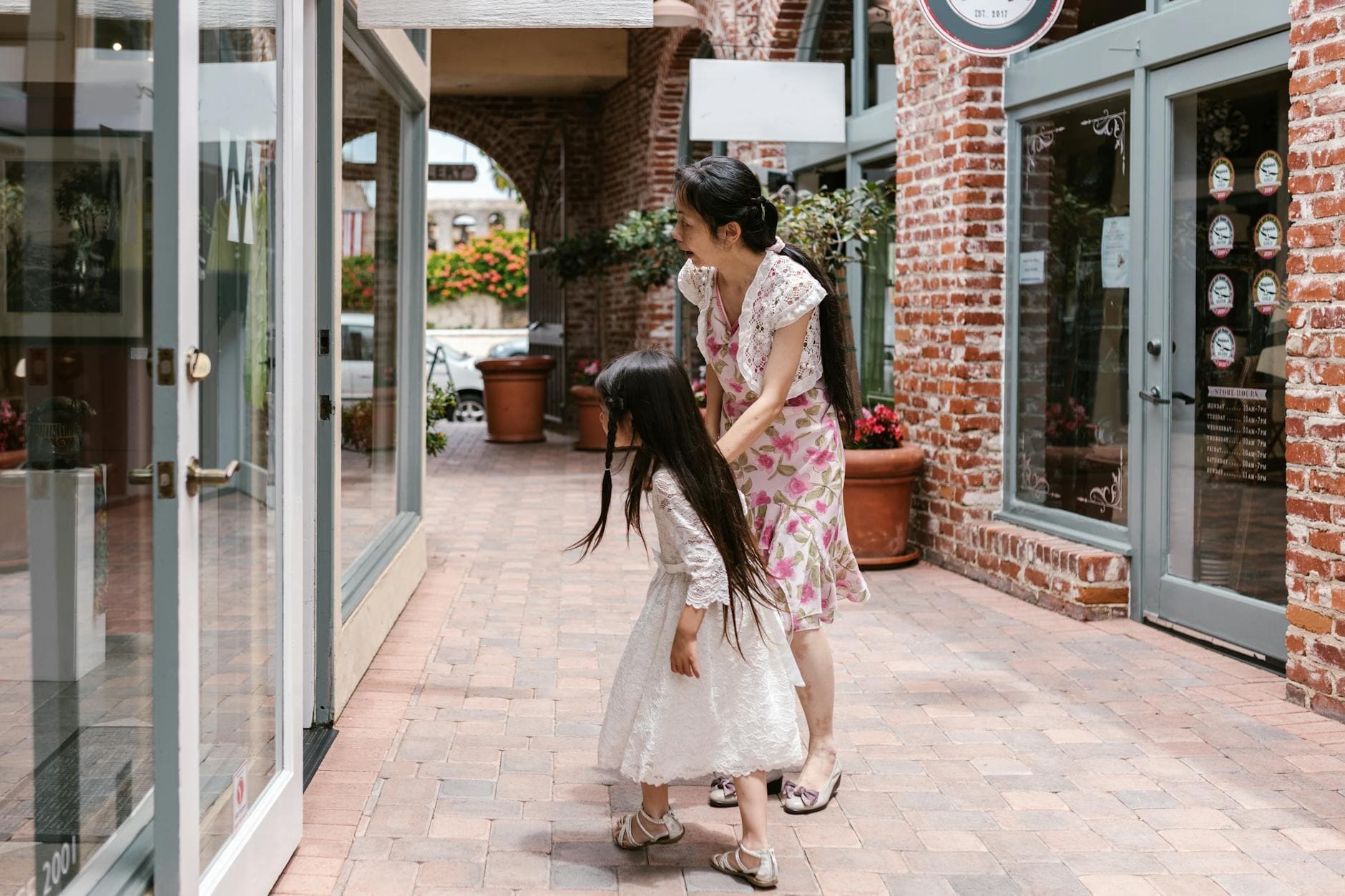 A mother and daughter window shopping in a charming urban brick-lined walkway.
