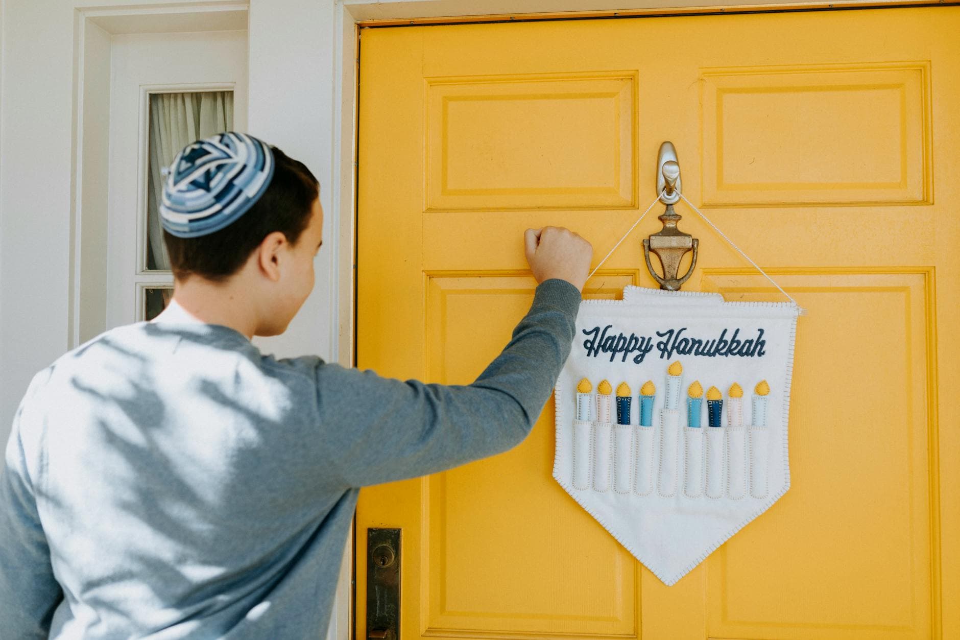 Boy wearing kippah knocks on a door decorated for Hanukkah with festive menorah banner.