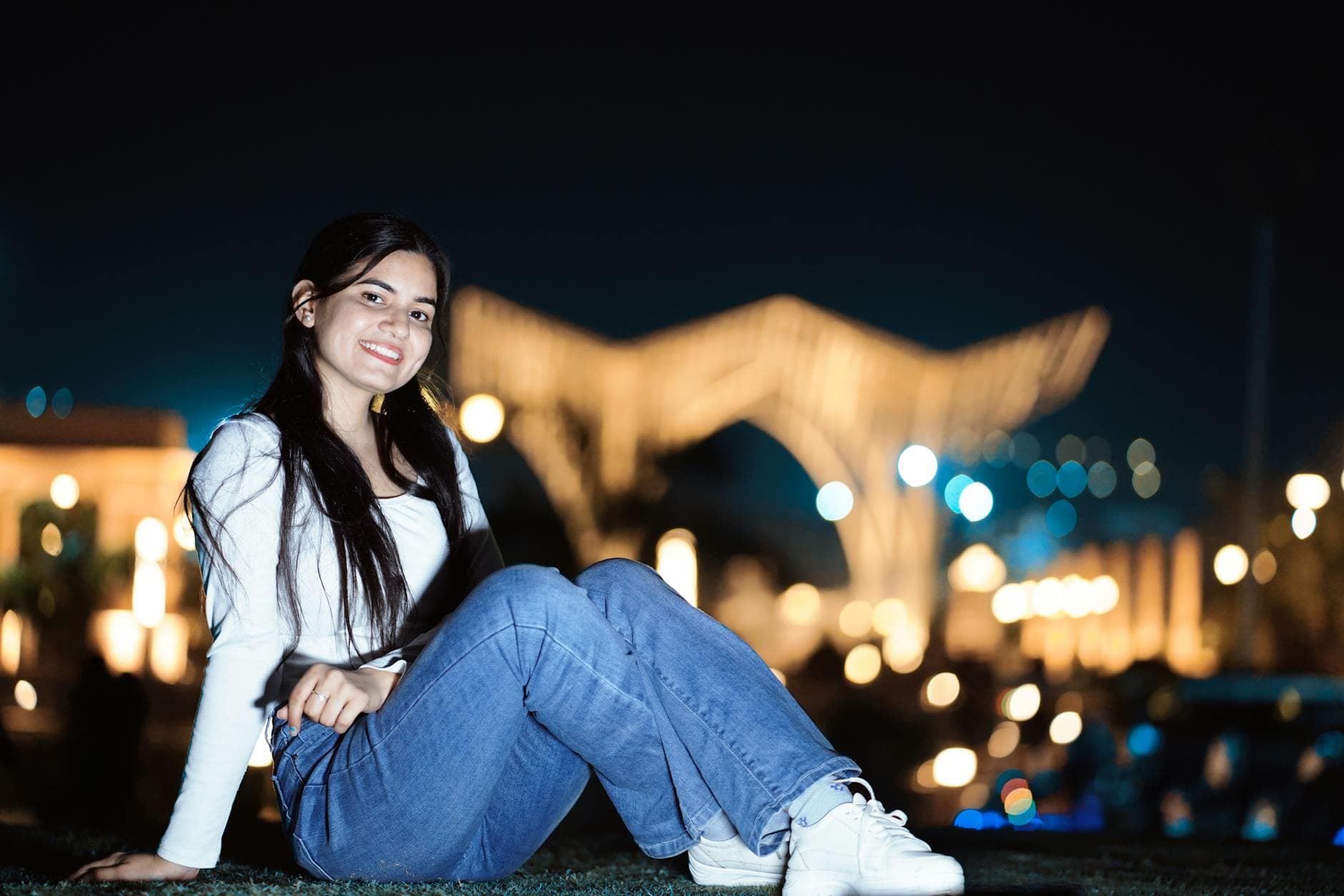 Young woman posing with a smile in an outdoor nighttime setting with blurred lights.
