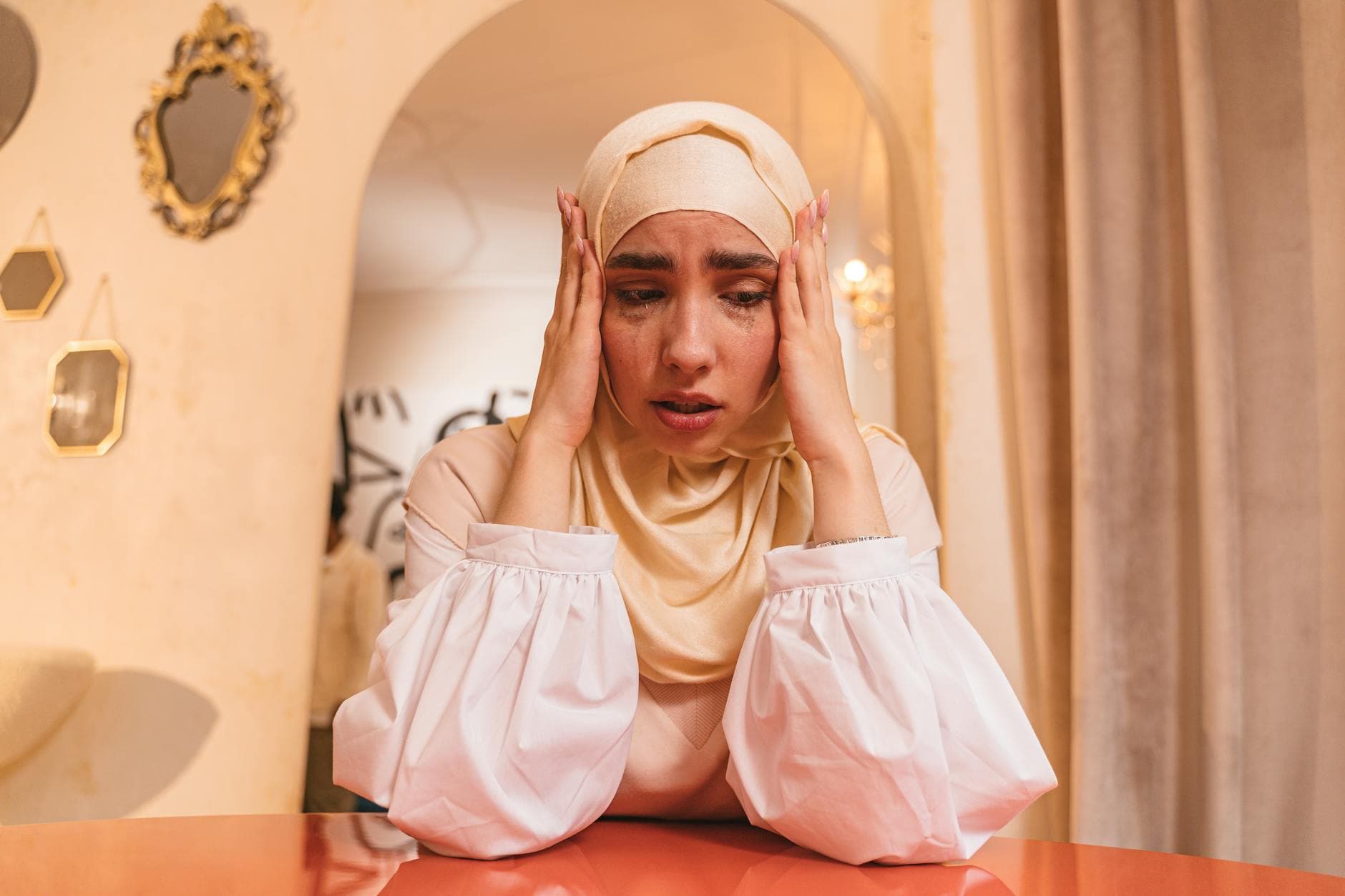 A woman in a hijab sitting at a table, expressing stress and emotion, with hands on her head.